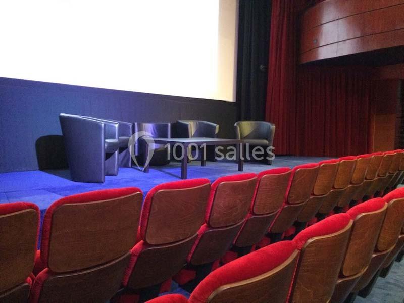 Salle de conférence avec rangées de sièges rouges, scène équipée de fauteuils noirs et d'une table basse.