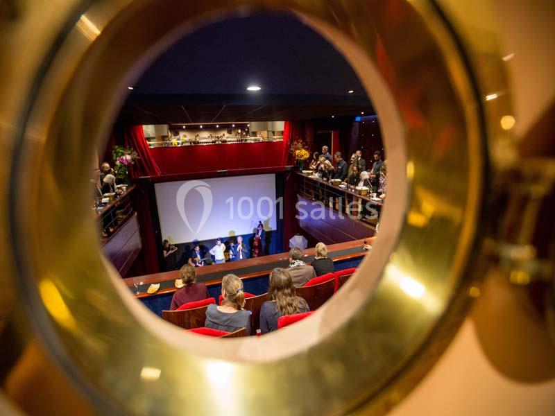 Vue à travers un hublot d'une salle de spectacle avec un écran, des spectateurs et des intervenants sur scène.