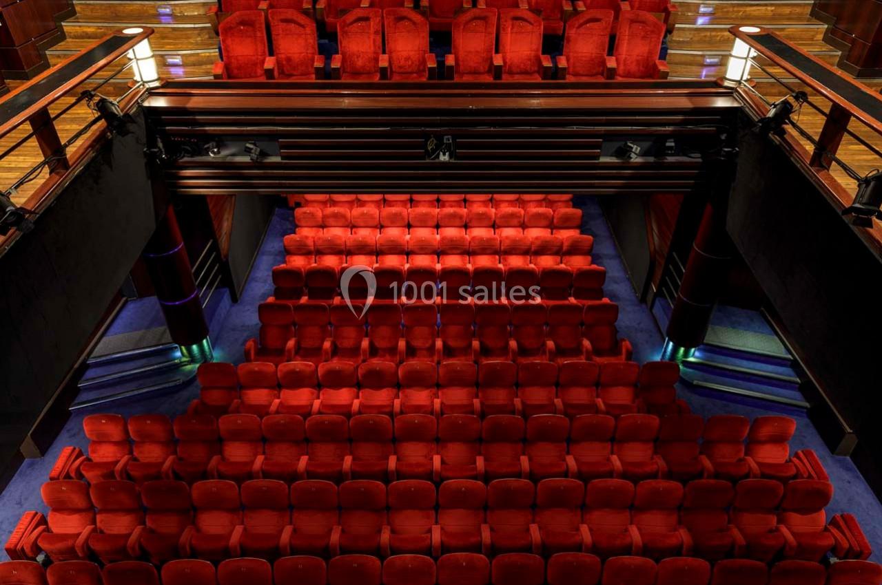 Salle de théâtre avec plusieurs rangées de sièges rouges vides et un balcon supérieur en bois.