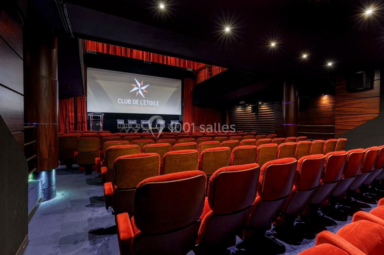 Salle de projection avec sièges rouges, écran géant affichant ’Club de l'Étoile’ et estrade équipée de chaises.