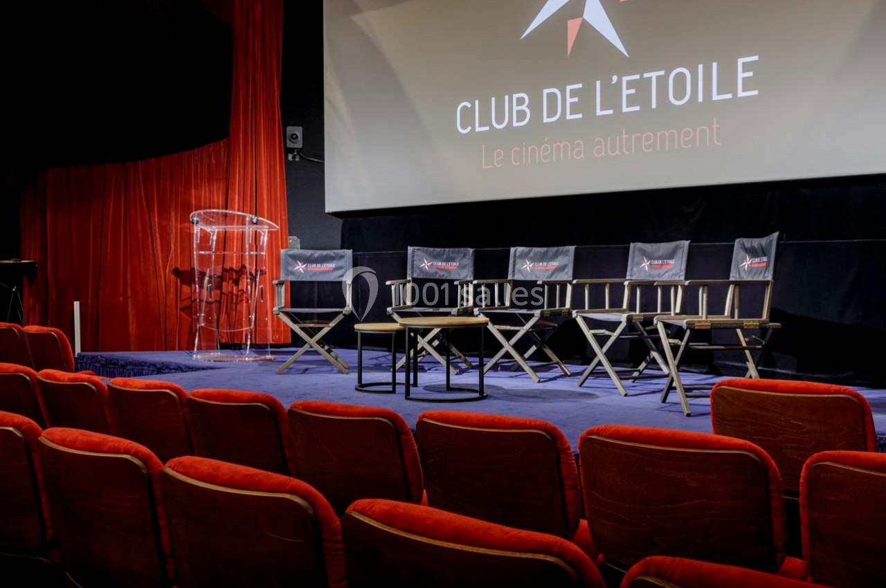 Salle de cinéma avec fauteuils rouges, estrade équipée de chaises pliantes et pupitre transparent devant un écran.