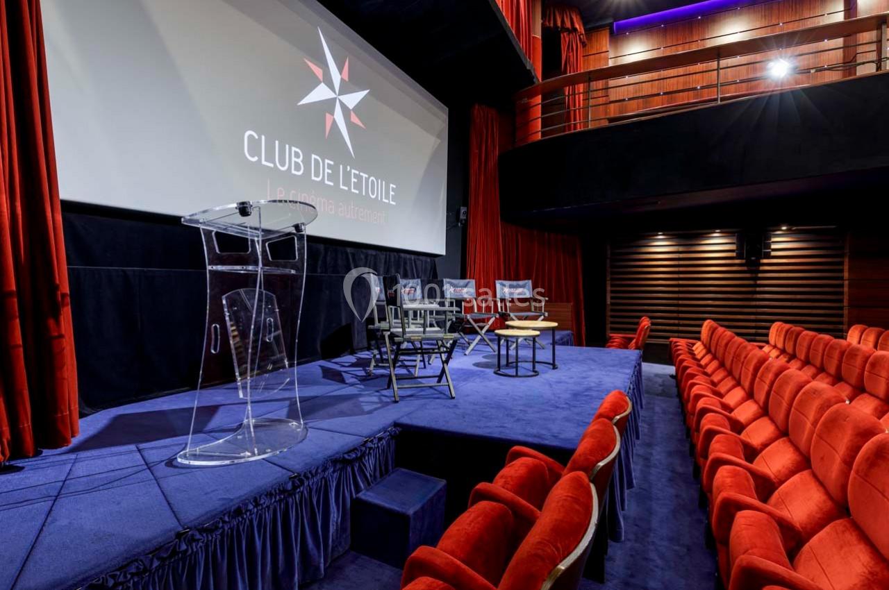 Salle de projection avec fauteuils rouges, scène équipée de chaises, table basse et pupitre transparent devant un écran.