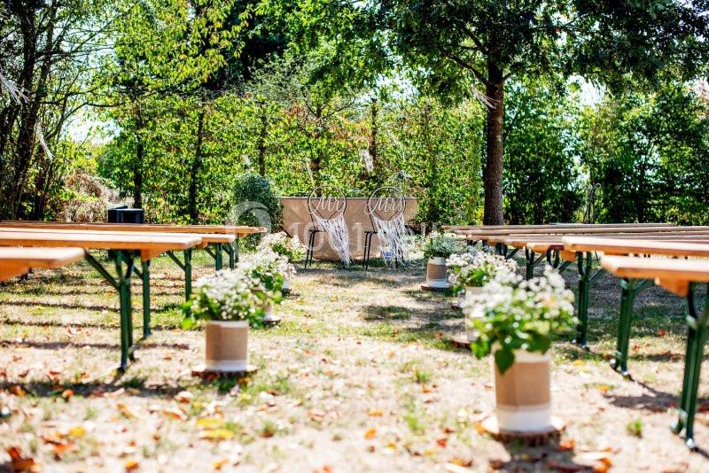 Chaises décorées et bancs en bois disposés en extérieur pour une cérémonie, entourés de verdure et de fleurs.