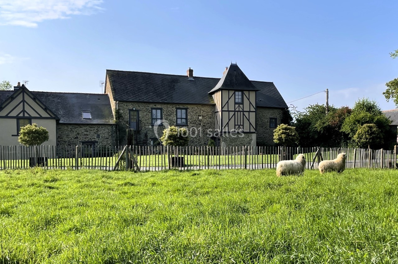 Maison en pierre avec colombages, entourée d'une clôture en bois, devant un pré où paissent deux moutons.