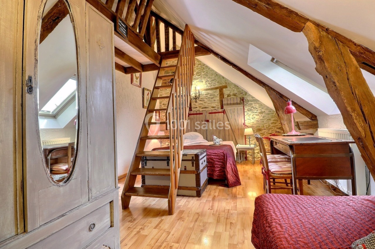 Chambre mansardée avec poutres apparentes, lit double, bureau en bois, coffre ancien et lumière naturelle.
