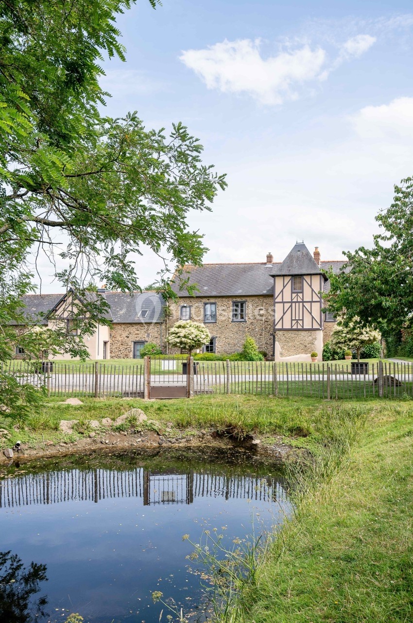 Façade d'une maison en pierre avec colombages, entourée de verdure, vue depuis un étang au premier plan.