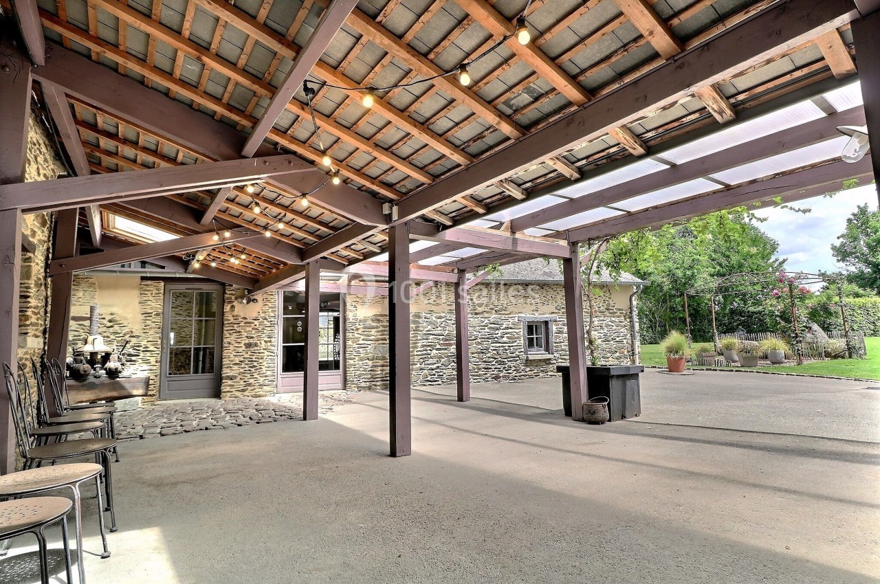 Terrasse couverte avec charpente en bois, éclairage suspendu et vue sur un bâtiment en pierre et un jardin.