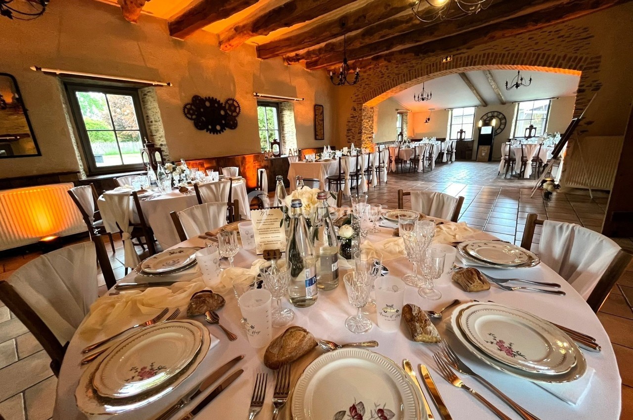 Salle de réception décorée avec des tables rondes dressées pour un repas, éclairage chaleureux et poutres apparentes.
