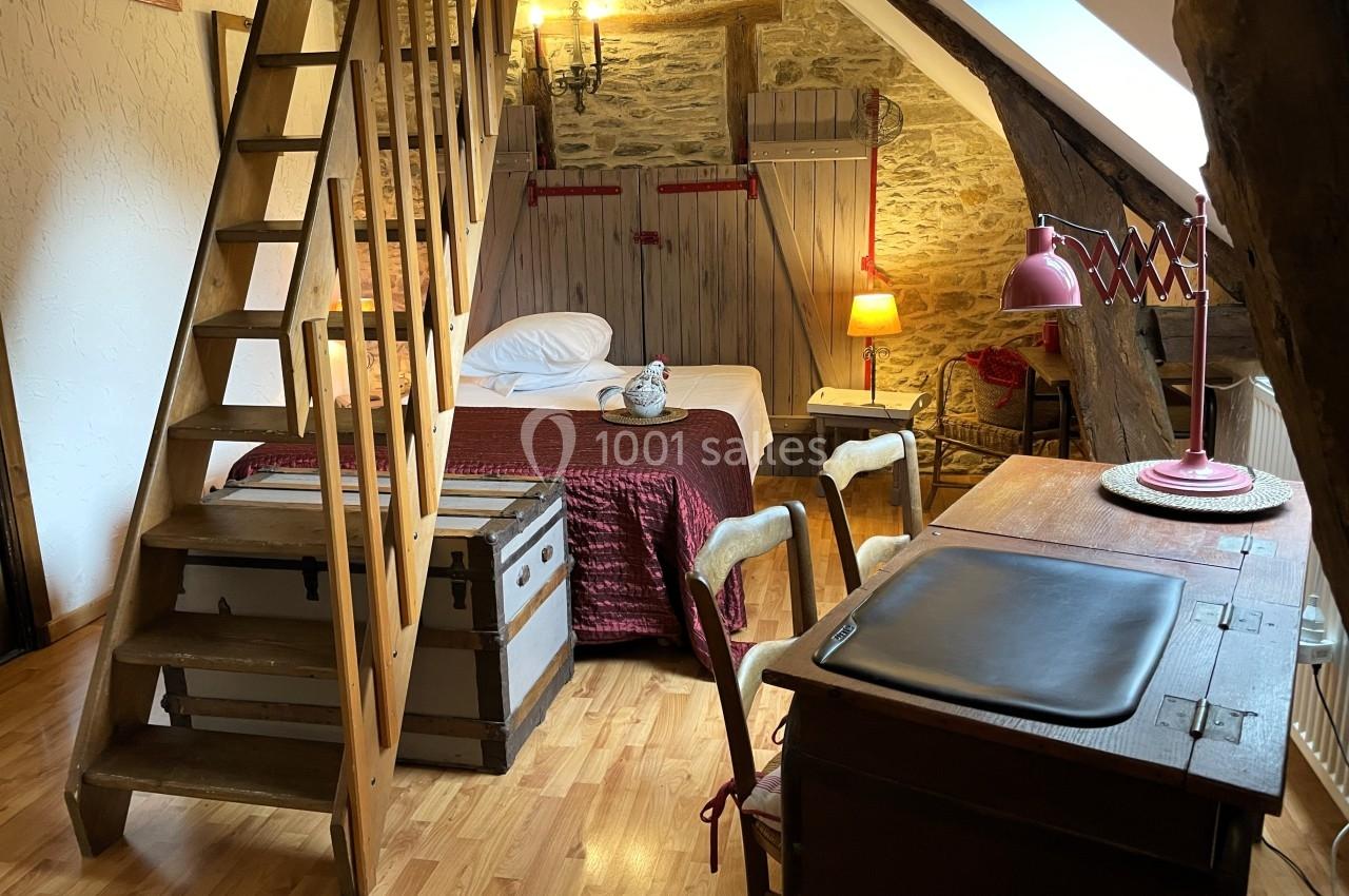 Chambre mansardée avec lit double, bureau en bois, malles anciennes et murs en pierre apparente.