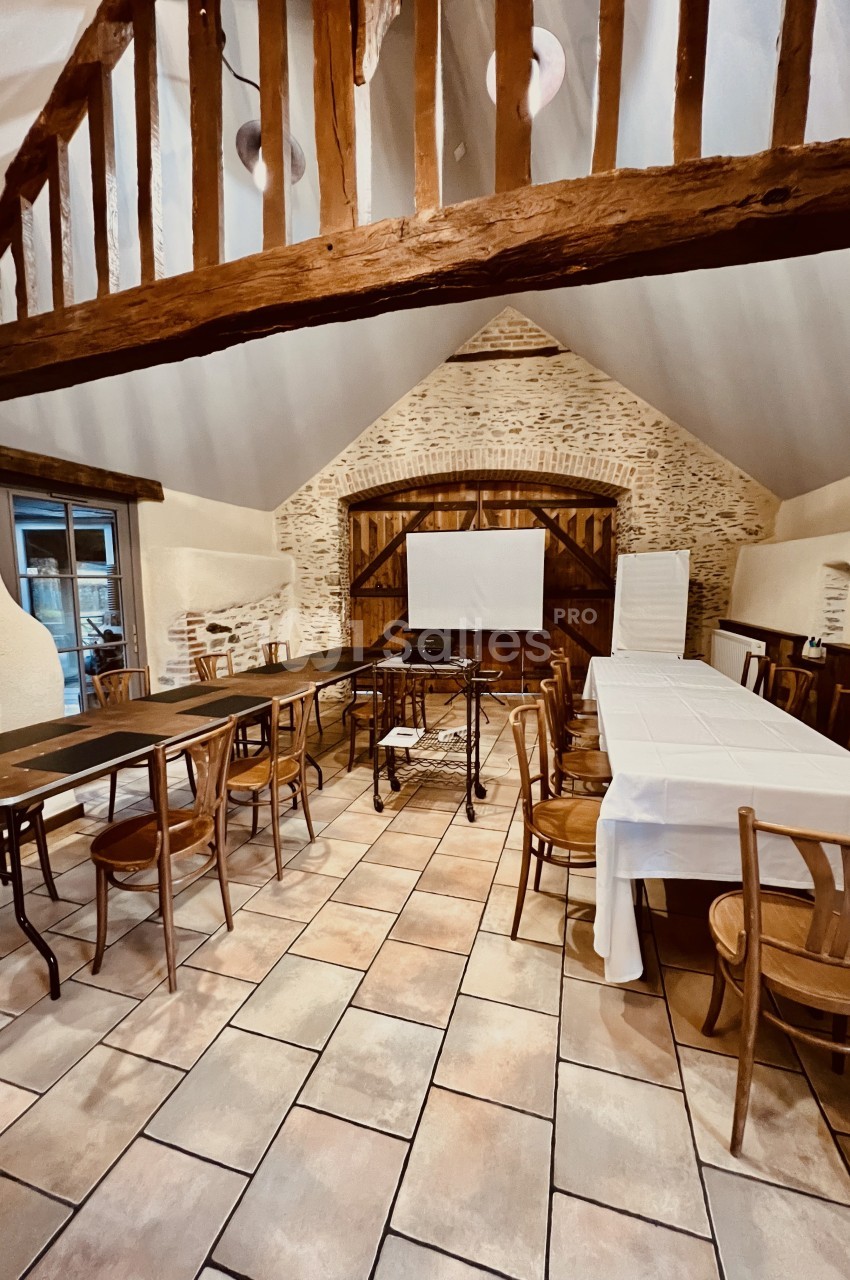 Salle rustique avec poutres apparentes, tables et chaises en bois disposées pour une réunion ou un événement.