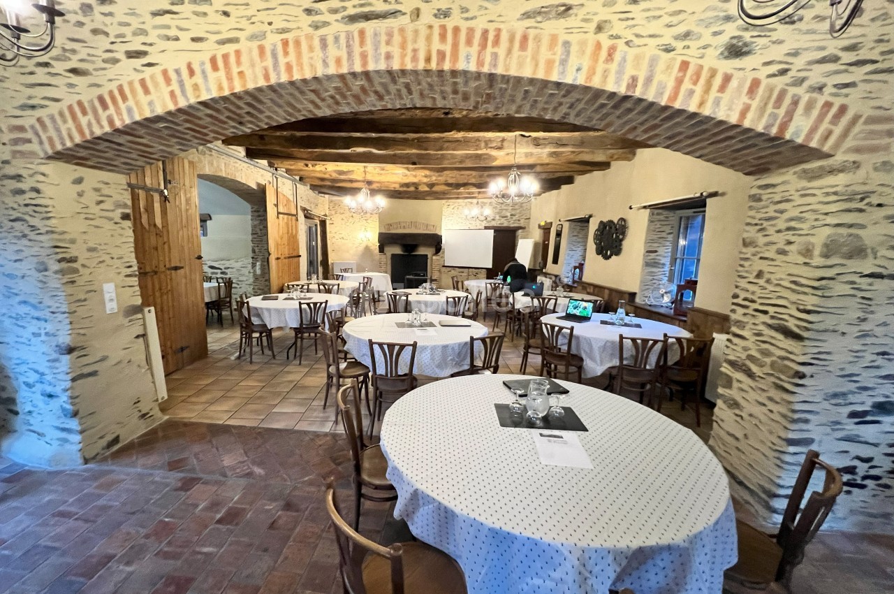 Salle en pierre avec des tables rondes dressées, chaises en bois, éclairage suspendu et cheminée en arrière-plan.