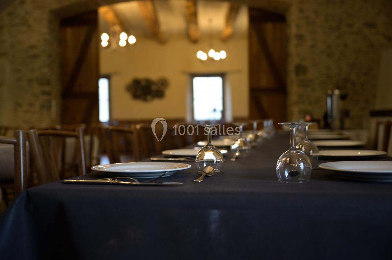 Table dressée avec assiettes, couverts et verres dans une salle en pierre avec poutres apparentes et éclairage chaleureux.