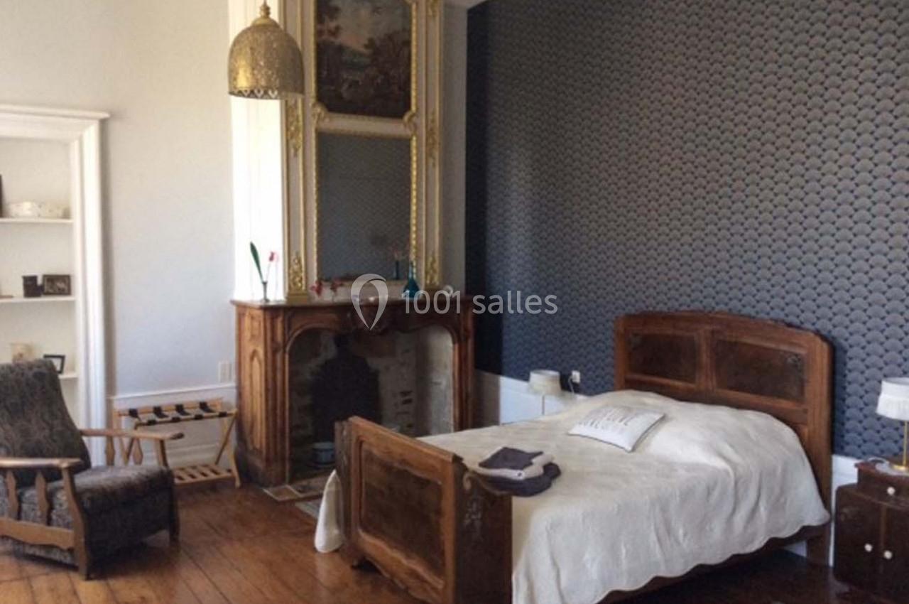 Chambre avec lit en bois, cheminée décorative, fauteuil et mur tapissé de motifs géométriques.