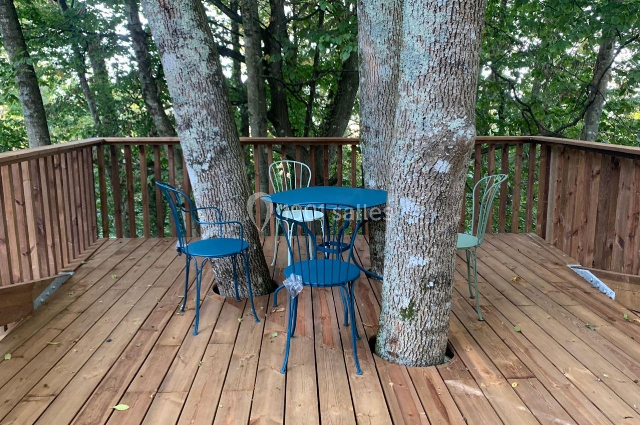 Terrasse en bois entourant des arbres, avec des chaises et une table en métal bleu disposées au centre.