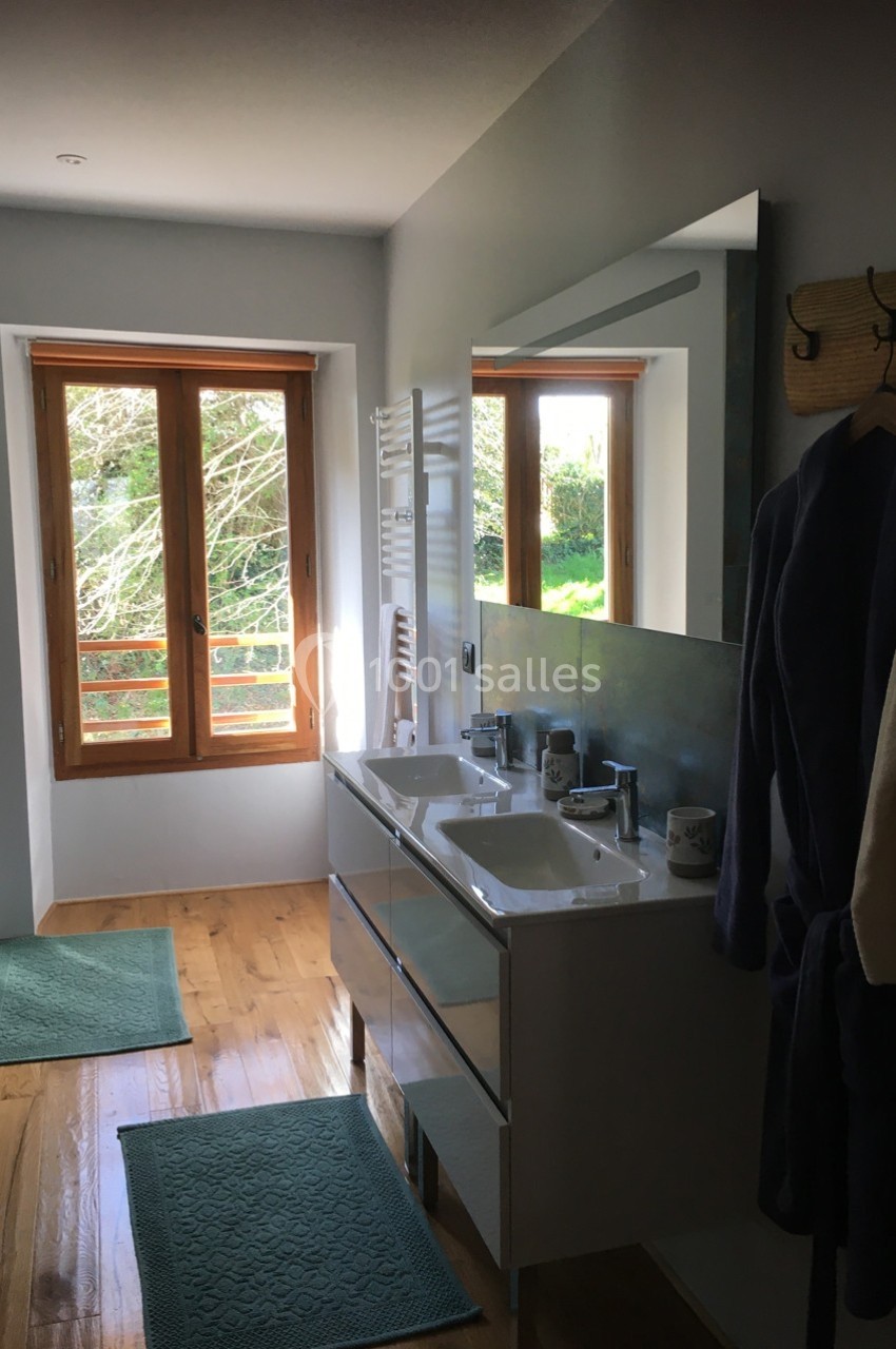 Salle de bain lumineuse avec double vasque, miroirs, tapis verts, parquet et fenêtre donnant sur un jardin.
