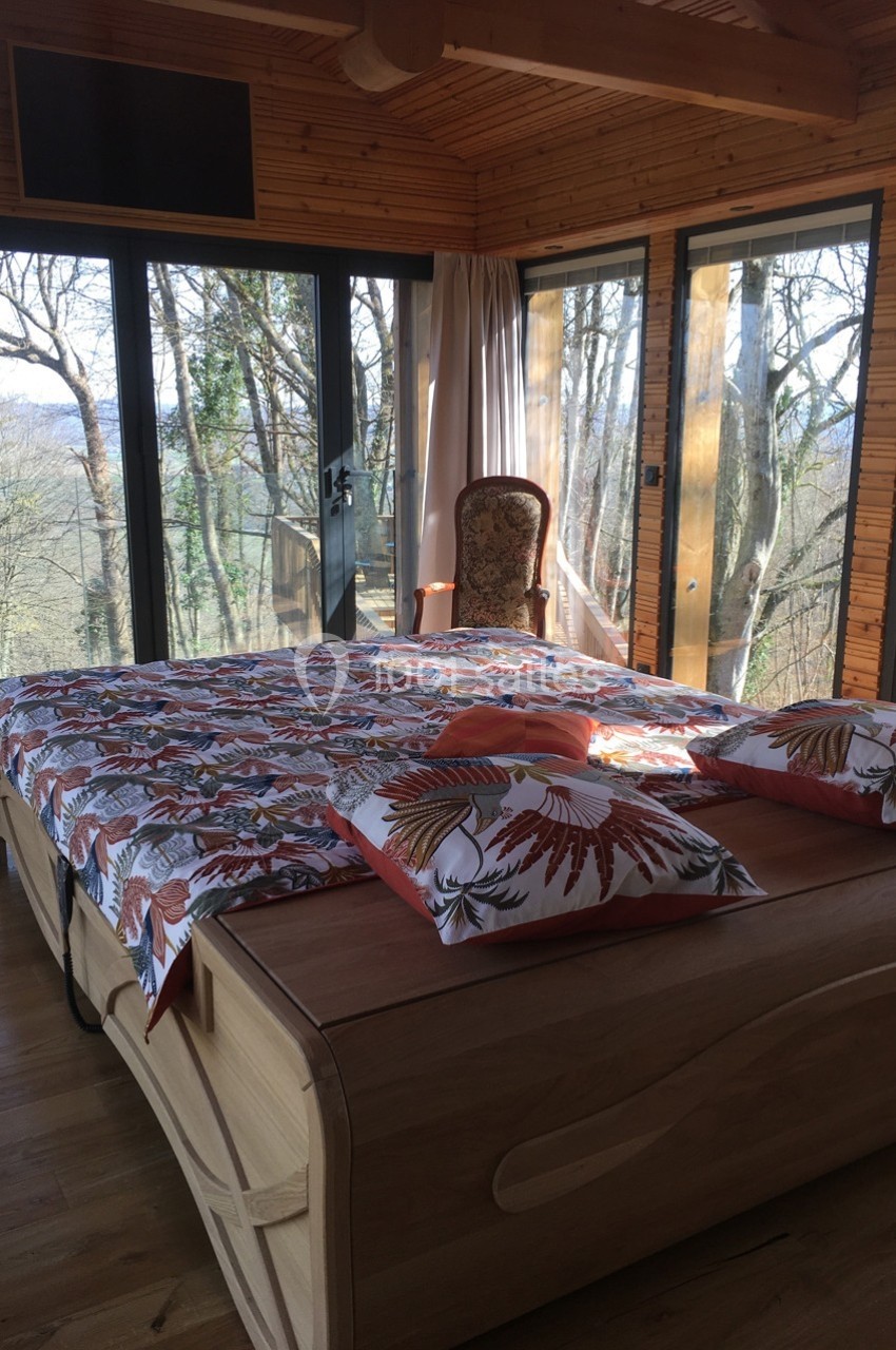 Chambre lumineuse avec lit en bois, grande baie vitrée donnant sur une forêt et fauteuil ancien près des rideaux.