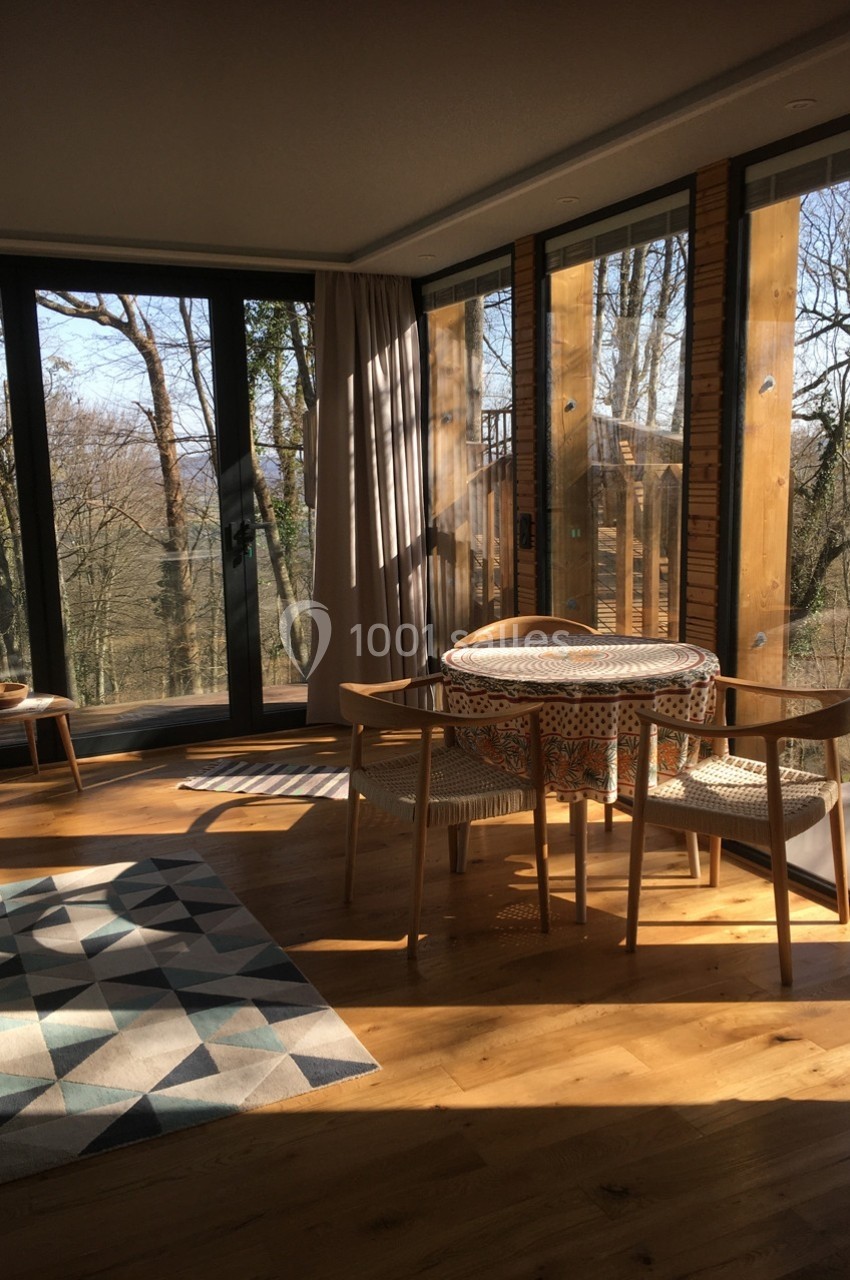 Salle à manger lumineuse avec table ronde, chaises en bois et grandes baies vitrées donnant sur une forêt.