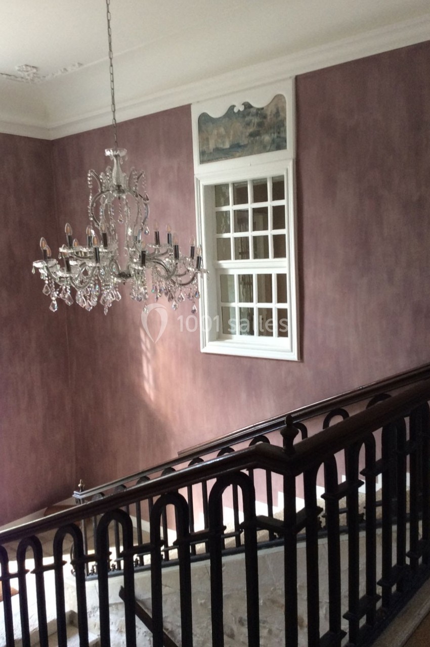 Escalier intérieur avec rampe en bois sombre, mur violet texturé, lustre en cristal et fenêtre blanche.