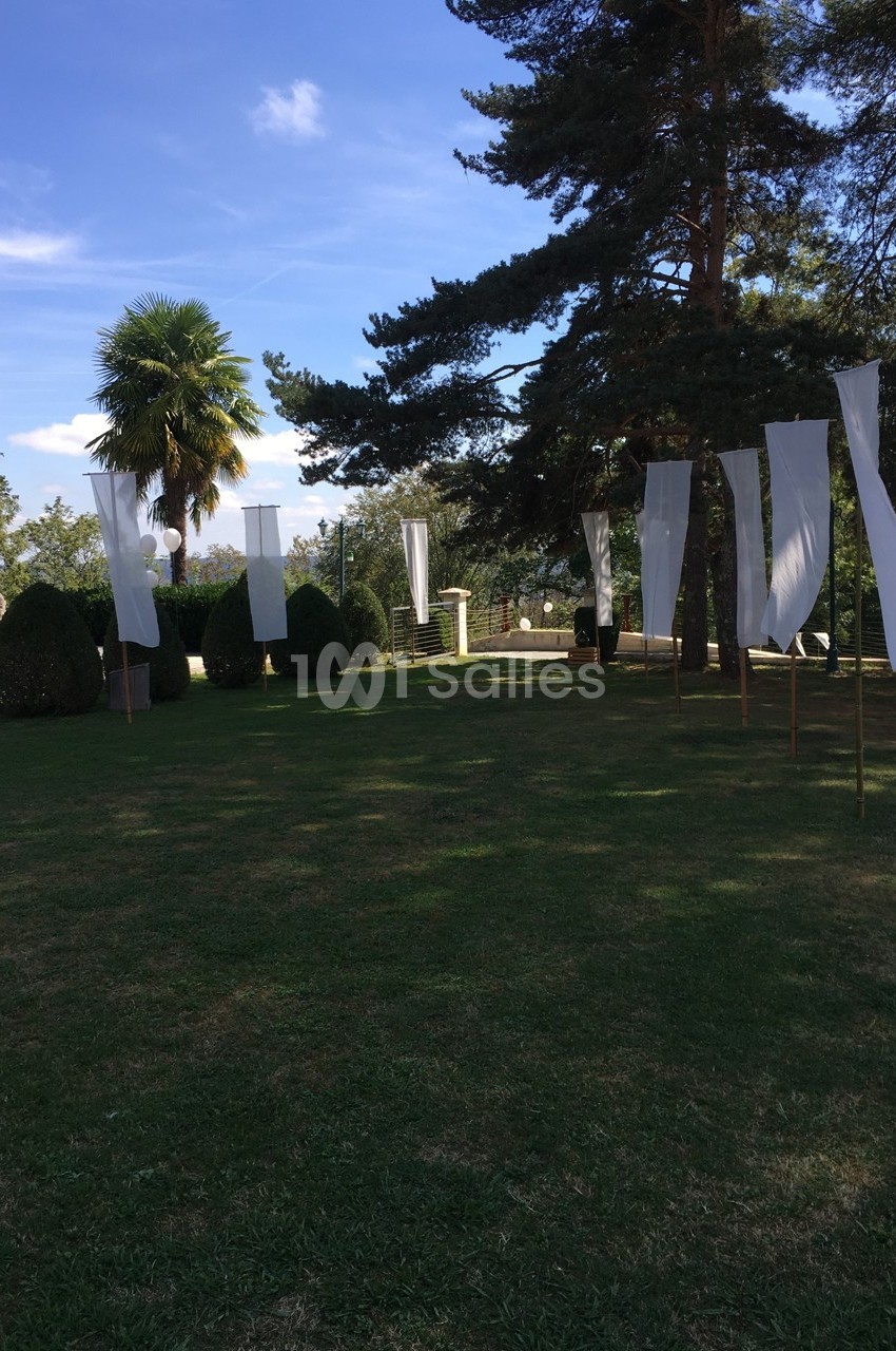 Des drapeaux blancs verticaux flottent dans un jardin herbeux entouré d'arbres sous un ciel bleu.
