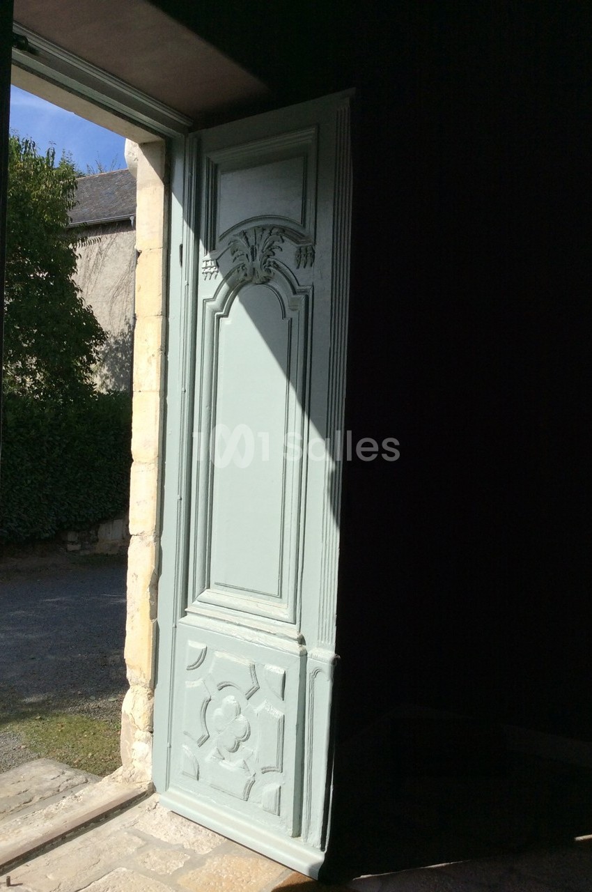 Porte en bois sculpté ouverte sur un extérieur ensoleillé avec un mur de pierre et des arbres en arrière-plan.