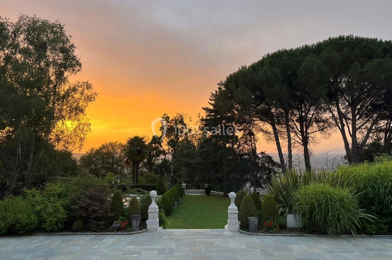 Coucher de soleil sur un jardin arboré avec une allée centrale bordée de verdure et des balustrades blanches.