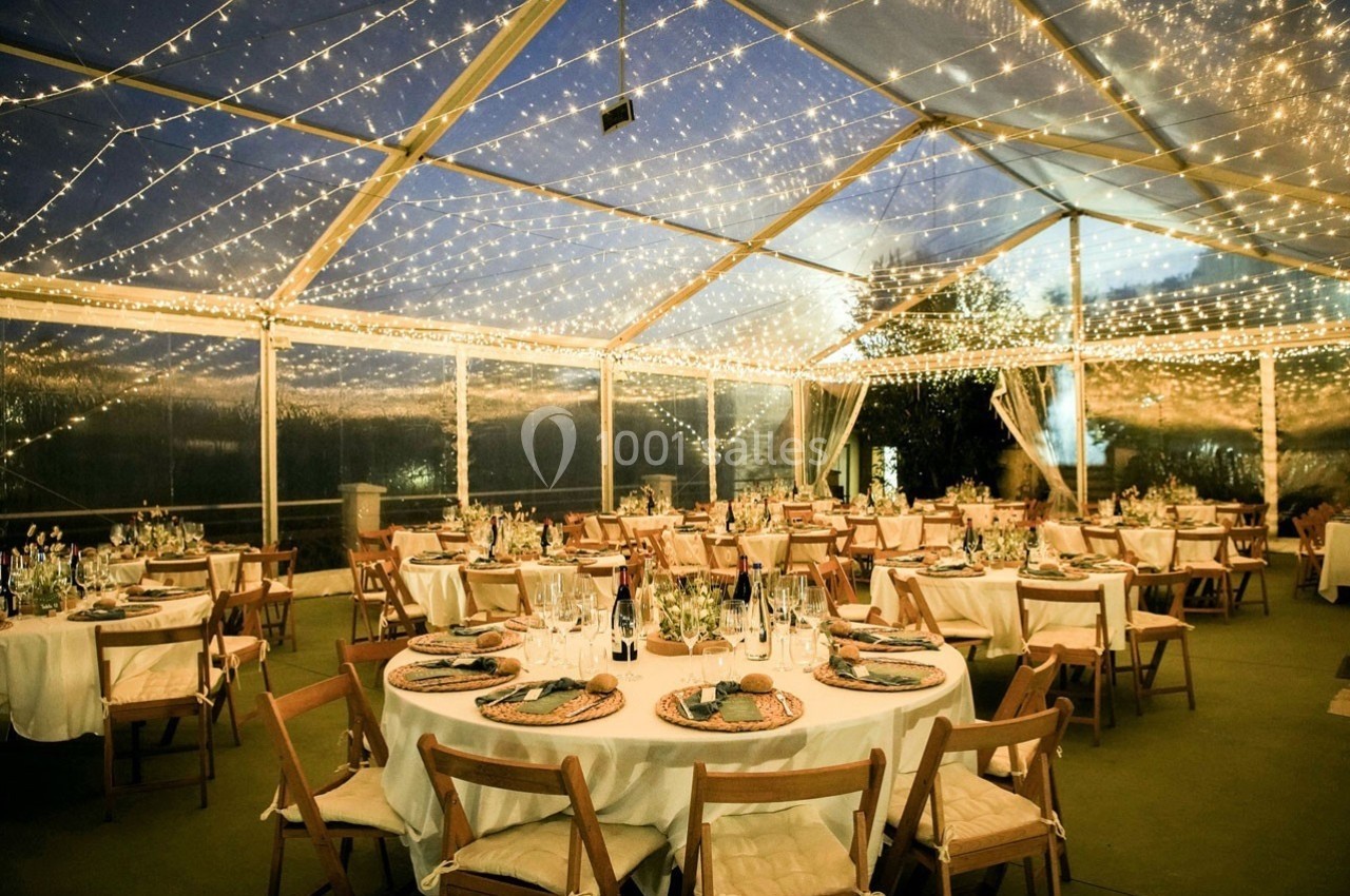 Salle de réception sous une tente transparente éclairée par des guirlandes lumineuses, avec tables rondes dressées.