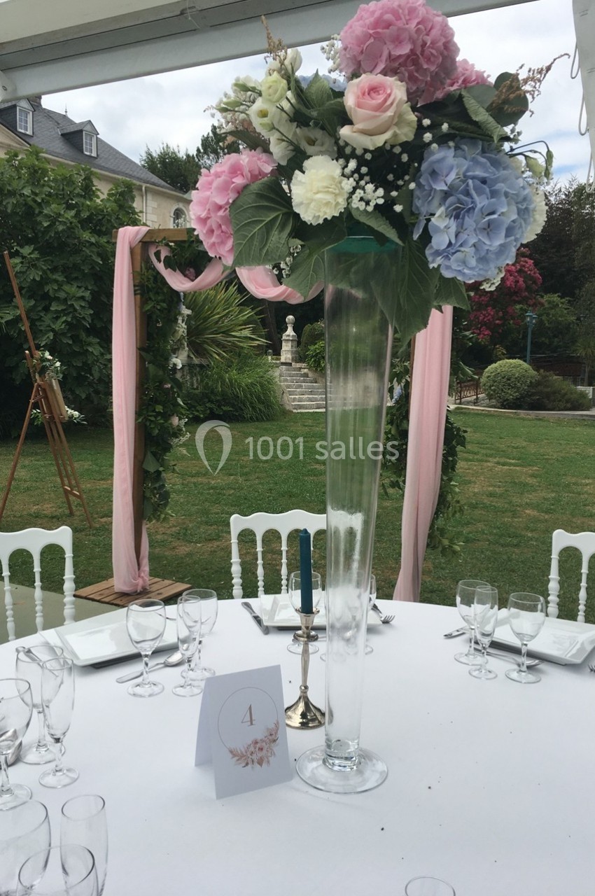 Centre de table floral avec roses et hortensias dans un vase haut, installé sur une table dressée en extérieur.