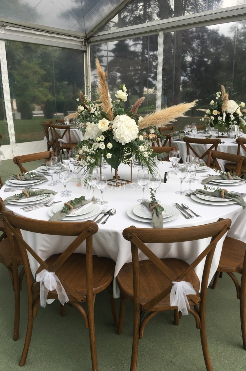 Table ronde décorée pour un événement, avec nappes blanches, chaises en bois et centre de table floral élégant.