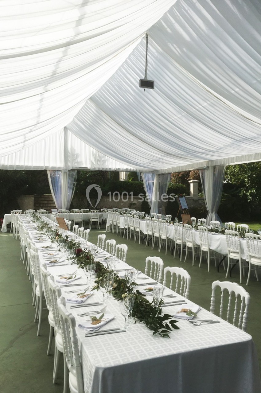 Salle de réception extérieure avec tables alignées, nappes blanches, chaises assorties et décoration végétale sous une tente.