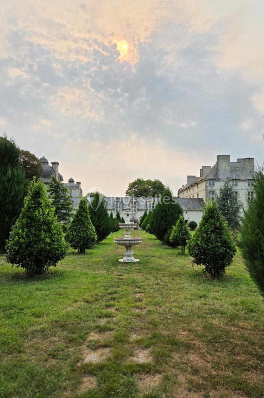 Allée bordée d'arbres menant à une fontaine en pierre, avec un château en arrière-plan sous un ciel nuageux.