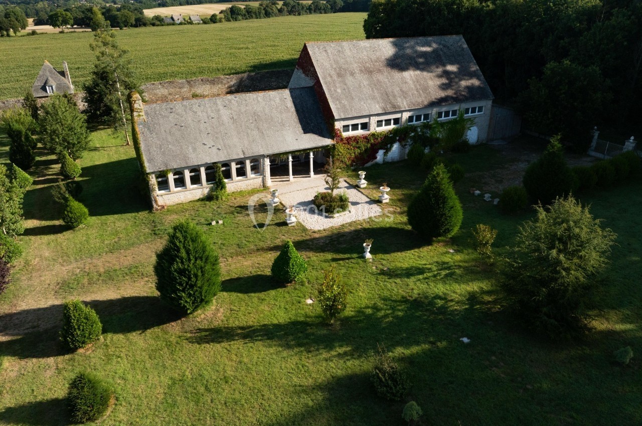 Vue aérienne d'un bâtiment rural entouré de pelouses, d'arbres et de champs, avec une forêt en arrière-plan.