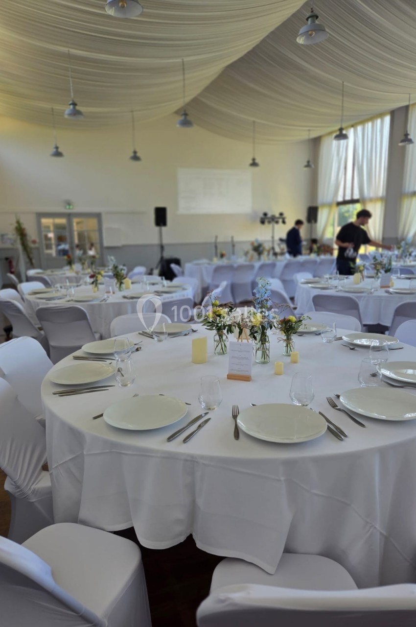 Salle de réception décorée avec des tables rondes dressées, nappes blanches et arrangements floraux.