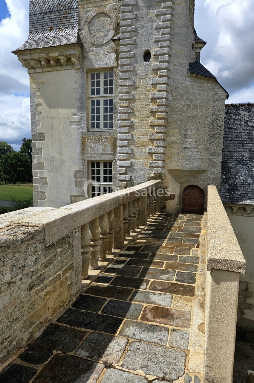 Passerelle en pierre bordée de balustrades menant à une porte en bois sur une tour en pierre ancienne.