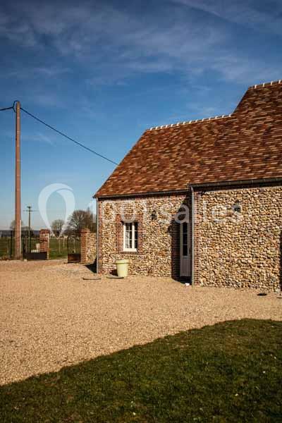 Maison en pierre avec toit en tuiles, située dans un environnement rural sous un ciel bleu dégagé.