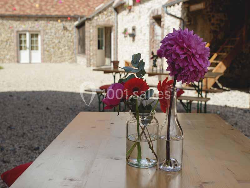 Deux vases avec des fleurs colorées posés sur une table en bois dans une cour pavée avec des bâtiments en arrière-plan.