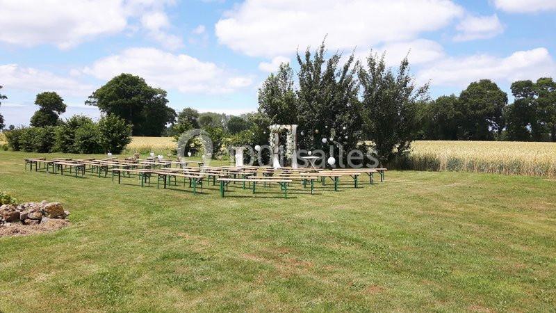 Location salle Saint-Pôtan (Côtes-d'Armor) - La Métairie de la Barre #9 Chaises et tables disposées en extérieur sur une pelouse, avec une arche décorative devant un champ et des arbres.