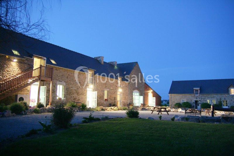 Location salle Saint-Pôtan (Côtes-d'Armor) - La Métairie de la Barre #7 Bâtiments en pierre éclairés de nuit, entourés de végétation et d'une cour avec des bancs en bois.