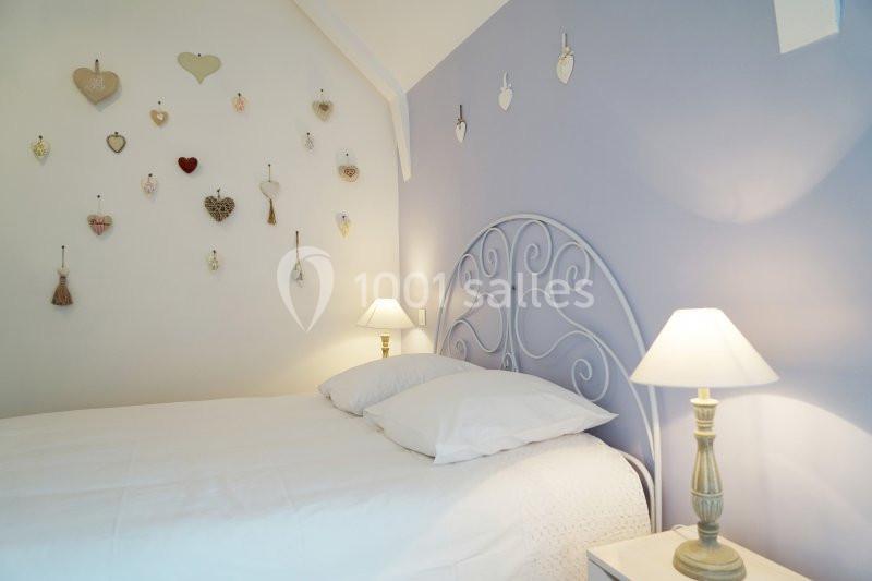 Location salle Saint-Pôtan (Côtes-d'Armor) - La Métairie de la Barre #18 Lit avec tête de lit en métal, draps blancs, mur violet décoré de cœurs suspendus, éclairé par deux lampes de chevet.