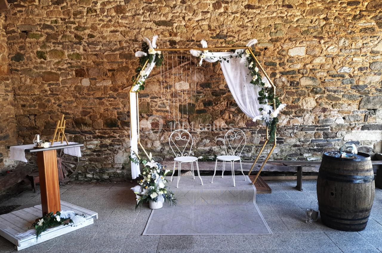 Location salle Saint-Pôtan (Côtes-d'Armor) - La Métairie de la Barre #4 Décor de cérémonie avec arche hexagonale ornée de fleurs blanches, deux chaises et un pupitre en bois devant un mur en…