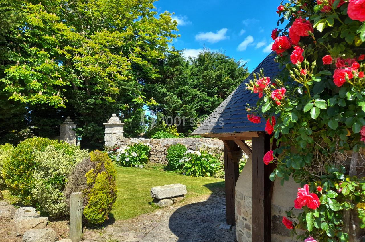 Location salle Saint-Pôtan (Côtes-d'Armor) - La Métairie de la Barre #12 Jardin fleuri avec rosiers rouges, pelouse, muret en pierre et arbres sous un ciel bleu ensoleillé.