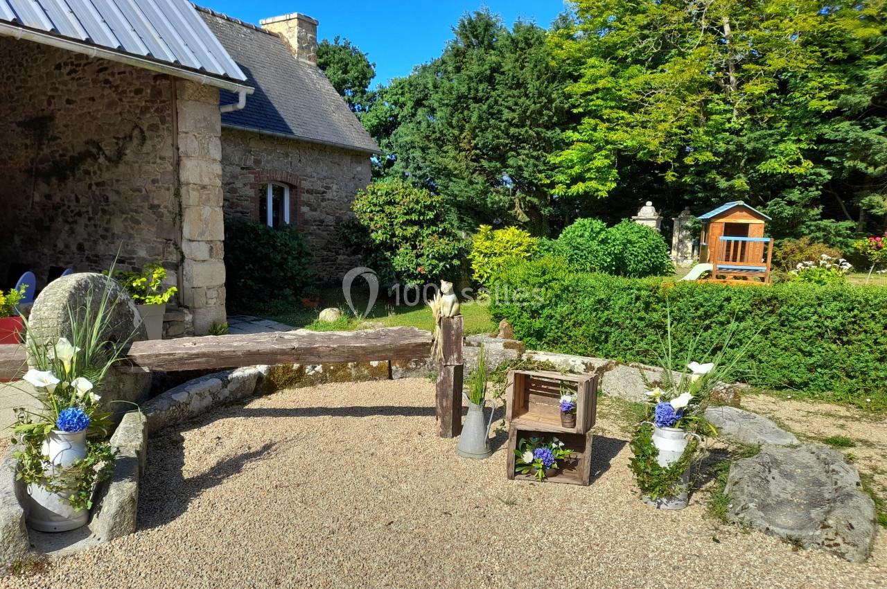 Location salle Saint-Pôtan (Côtes-d'Armor) - La Métairie de la Barre #8 Cour extérieure avec décor champêtre, comprenant des fleurs, des caisses en bois et une petite maison en arrière-plan.