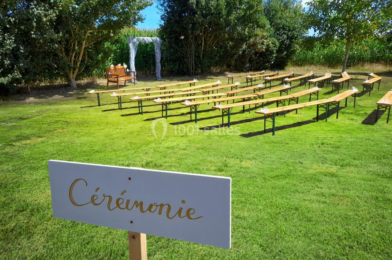 Location salle Saint-Pôtan (Côtes-d'Armor) - La Métairie de la Barre #2 Bancs en bois alignés en extérieur pour une cérémonie, avec une arche décorative et un panneau ’Cérémonie’.