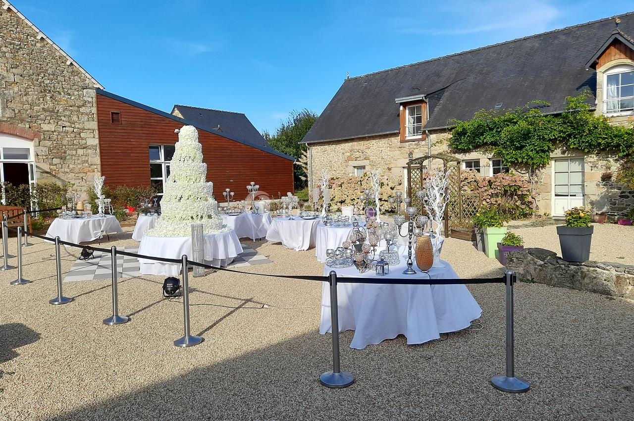 Location salle Saint-Pôtan (Côtes-d'Armor) - La Métairie de la Barre #3 Cour extérieure avec tables décorées pour un événement, pièce montée blanche au centre, entourée de bâtiments en pierre.