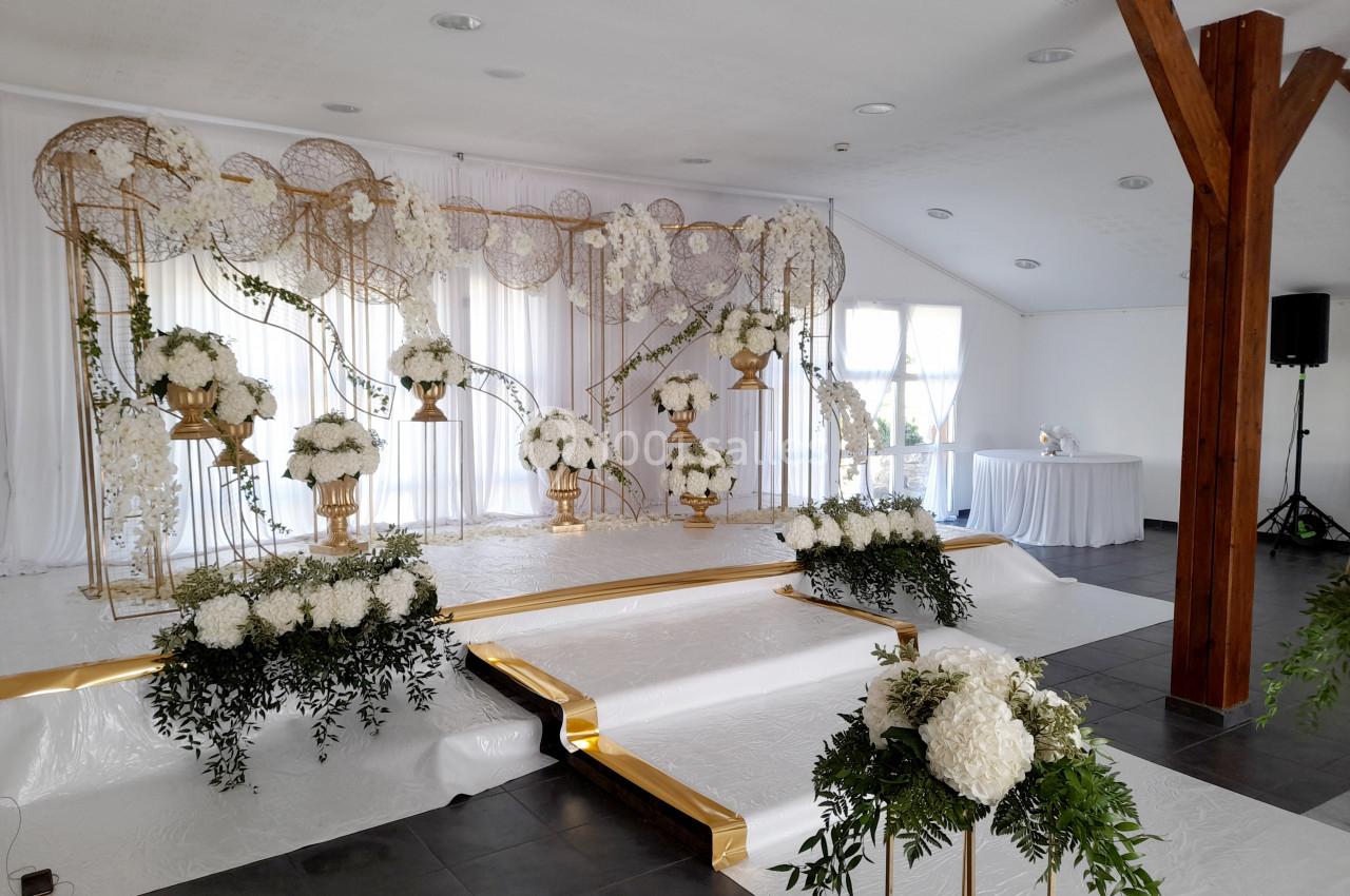 Location salle Saint-Pôtan (Côtes-d'Armor) - La Métairie de la Barre #10 Décoration de scène de mariage avec fleurs blanches, arches dorées et rideaux blancs dans une salle lumineuse.