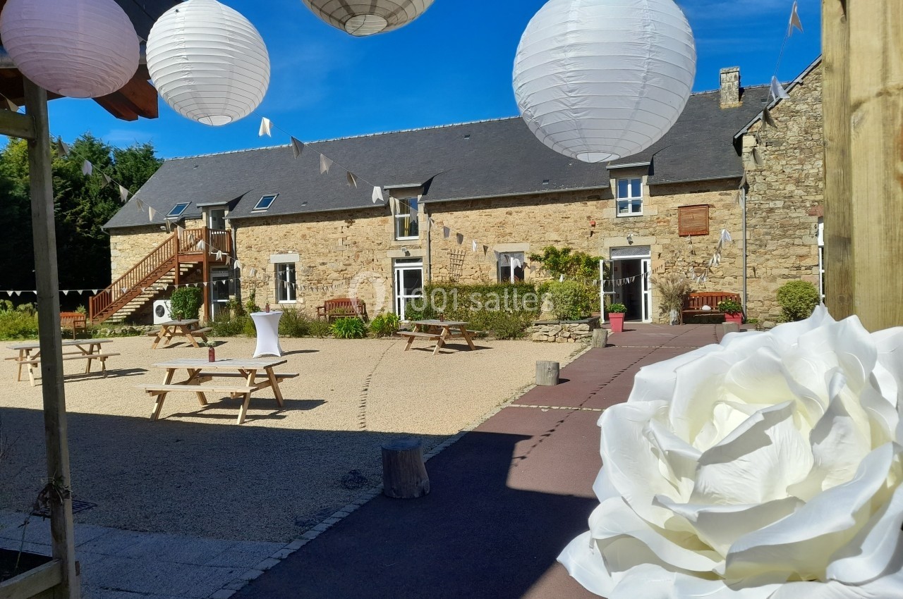 Location salle Saint-Pôtan (Côtes-d'Armor) - La Métairie de la Barre #23 Cour d'une bâtisse en pierre avec lanternes blanches suspendues, tables en bois et grande rose blanche au premier plan.