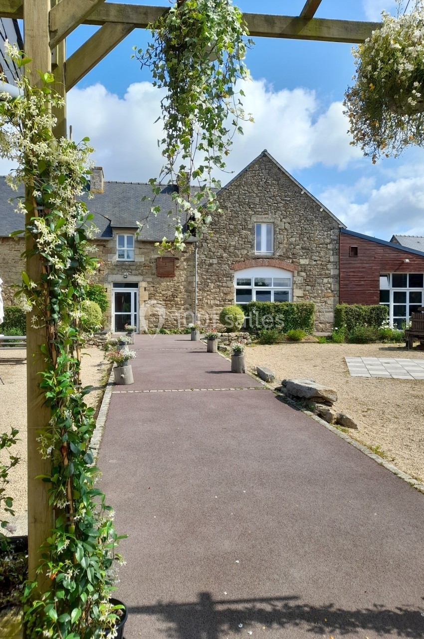 Location salle Saint-Pôtan (Côtes-d'Armor) - La Métairie de la Barre #26 Allée pavée bordée de plantes menant à une bâtisse en pierre sous un ciel partiellement nuageux.