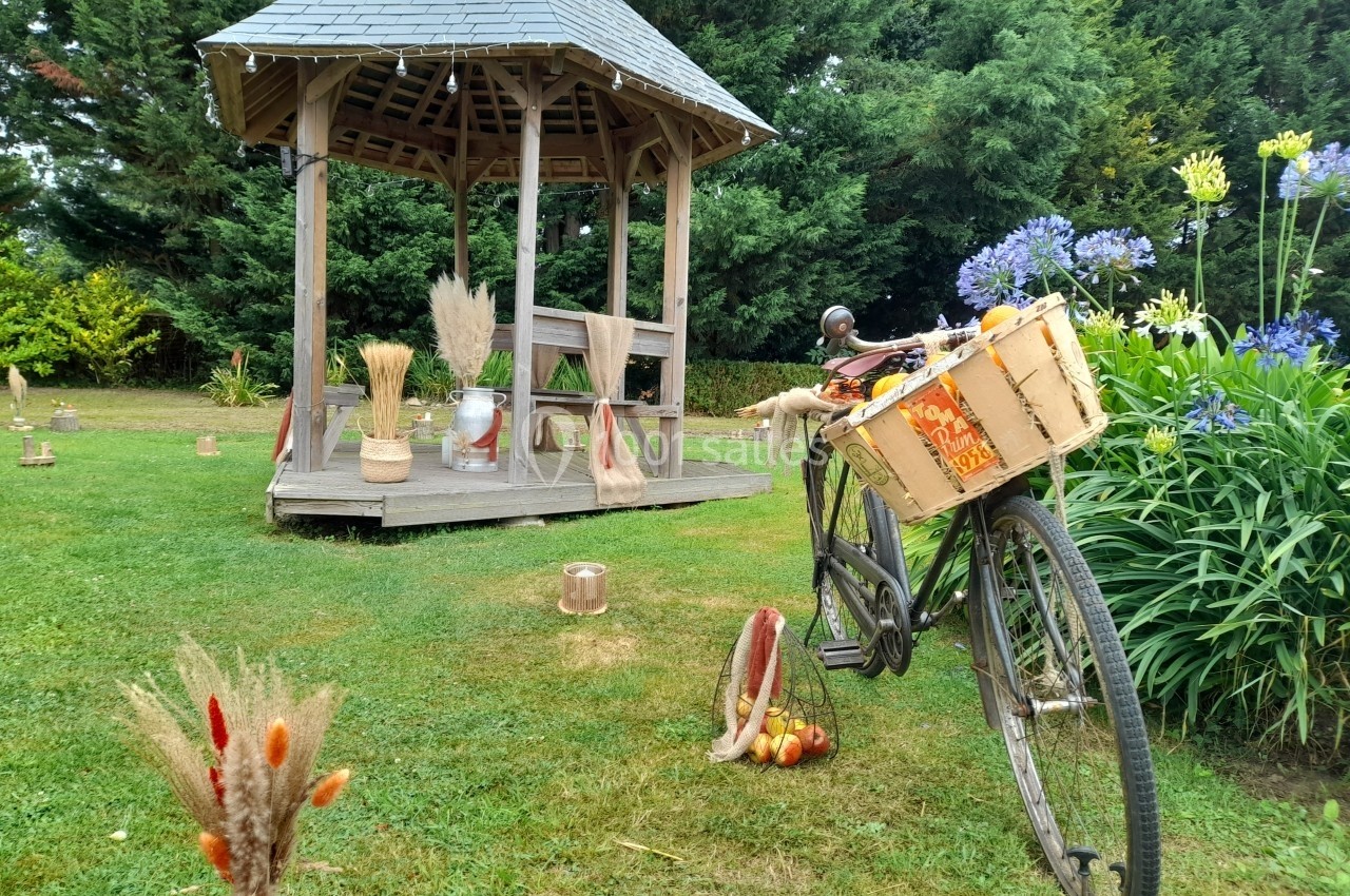 Location salle Saint-Pôtan (Côtes-d'Armor) - La Métairie de la Barre #27 Un vélo ancien avec un panier rempli de pommes est posé près d'un kiosque en bois dans un jardin verdoyant.