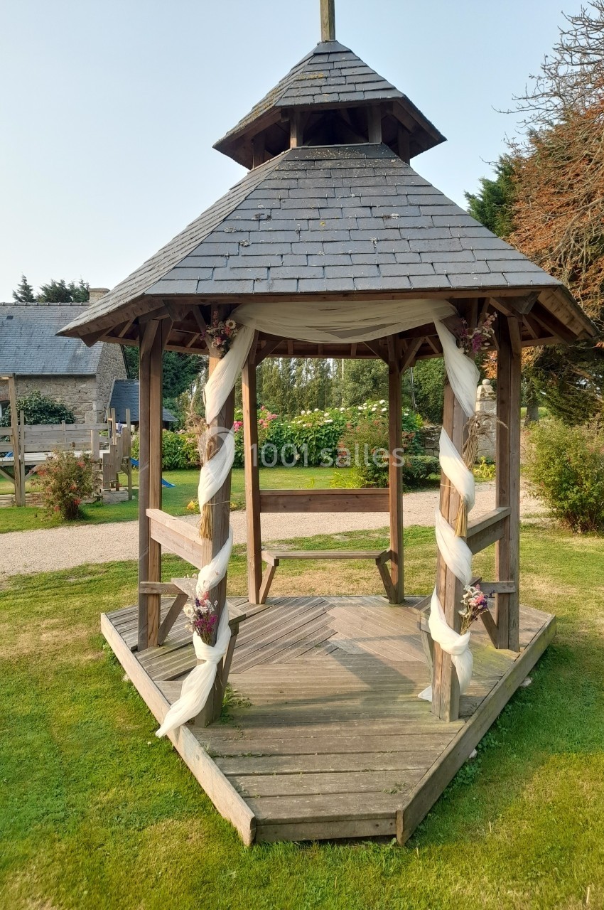 Location salle Saint-Pôtan (Côtes-d'Armor) - La Métairie de la Barre #31 Kiosque en bois décoré de voiles blancs et de fleurs, situé sur une pelouse dans un jardin.
