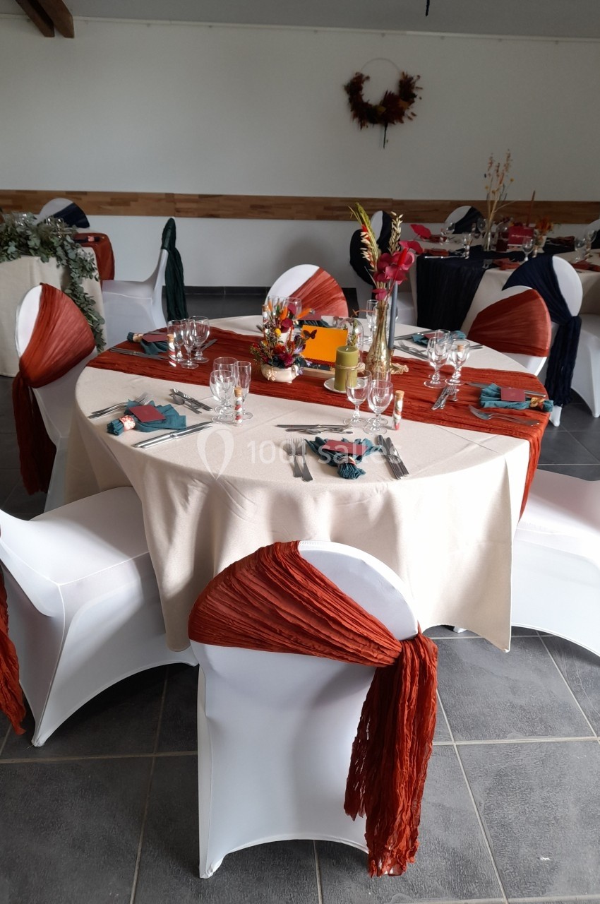 Location salle Saint-Pôtan (Côtes-d'Armor) - La Métairie de la Barre #32 Table ronde décorée pour un événement, avec nappes beige, chaises blanches ornées de tissu orange et centre de table floral.