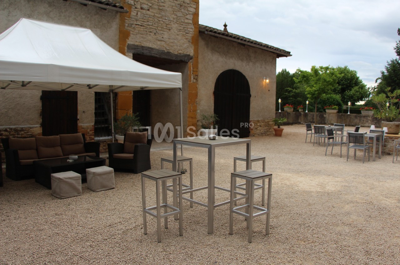 Mobilier de jardin avec tables hautes et canapés sous un barnum, installé dans une cour en gravier devant un bâtiment en…